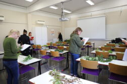 Luokkahuone. Pöydillä kasveja. Opiskelijat kiertämässä luokassa paperien ja kynien kanssa.