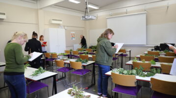 Luokkahuone. Pöydillä kasveja. Opiskelijat kiertämässä luokassa paperien ja kynien kanssa.
