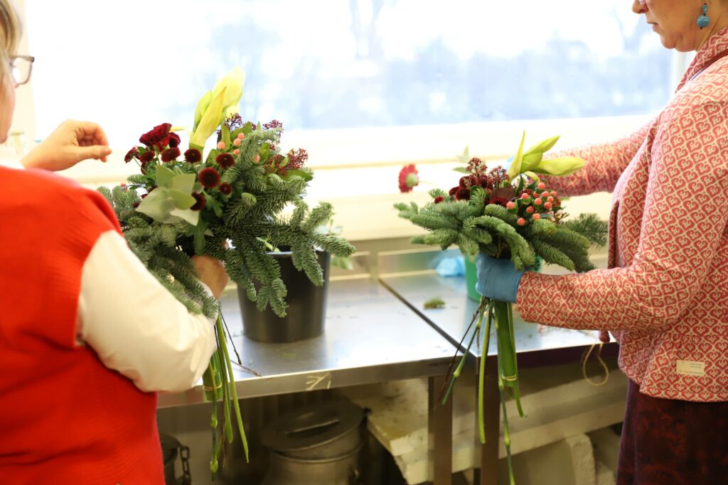 Kaksi naista näkyy osittain kuvassa. Valmistavat kukkakimppuja käsissään.