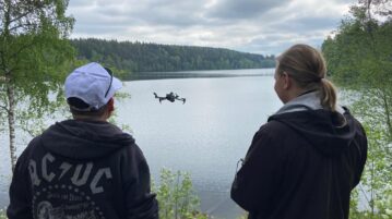 kaksi miestä lennättää dronea järven rannalla