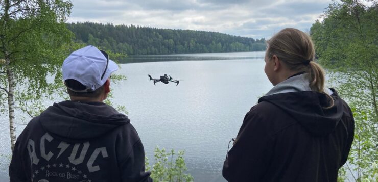 kaksi miestä lennättää dronea järven rannalla