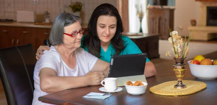 Hoitaja ja seniori-ikäinen henkilö tarkastelevat pöydän ääressä tablettitietokonetta