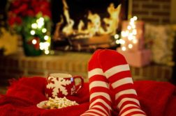Kuvan taustalla näkyy takka, jossa palaa tuli, takan vieressä loistavat jouluvalot. Edustalla näemme jalat punavalkoisissa sukissa. Jalkojen alla on punainen torkkupeite, jonka päälle on asetettu punavalkoinen muki ja lautasellinen joulupiparkakkuja.