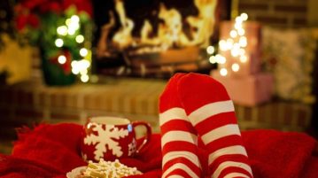 Kuvan taustalla näkyy takka, jossa palaa tuli, takan vieressä loistavat jouluvalot. Edustalla näemme jalat punavalkoisissa sukissa. Jalkojen alla on punainen torkkupeite, jonka päälle on asetettu punavalkoinen muki ja lautasellinen joulupiparkakkuja.